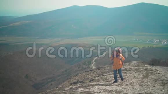 一个穿着黄色夹克蓝色牛仔裤和带着一个大型旅游背包的帽子的人穿过山脉向远处望去视频的预览图