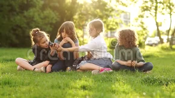孩子和小玩意同学们坐在公园的草地上互相递上一部智能手机日落视频的预览图