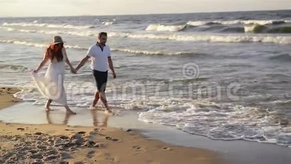 沙滩上恋爱的年轻夫妇穿着白色衣服的漂亮夫妇情人牵手拥抱视频的预览图
