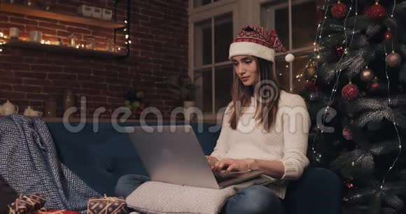 一位身穿圣诞帽坐在家中圣诞树的美丽年轻女子用手提电脑聊天视频的预览图