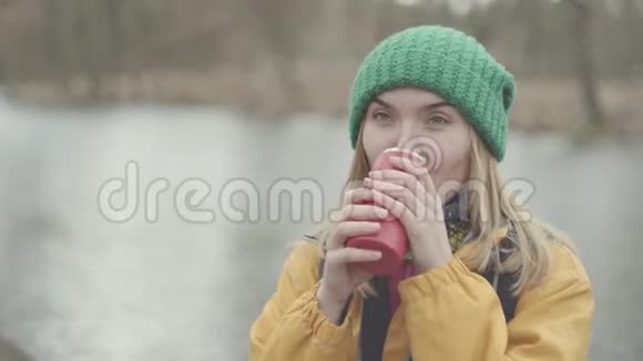 公园里戴着绿帽子穿着黄色外套从热水瓶里喝茶或喝咖啡的漂亮年轻女子的画像视频的预览图