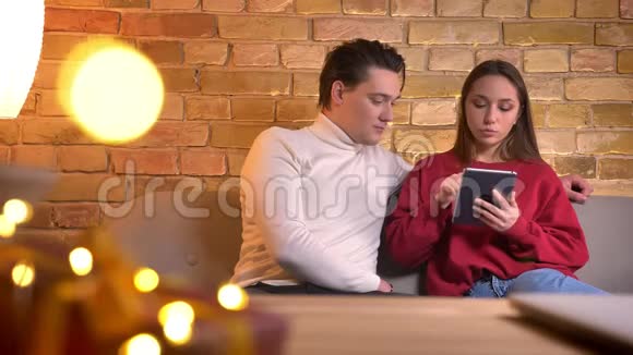 在家庭氛围中年轻的白种人朋友坐在沙发上专注地与平板电脑合作特写照片视频的预览图