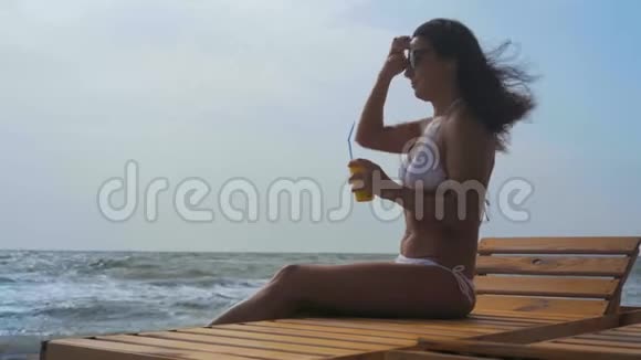 穿着白色泳衣的漂亮女人手里拿着鸡尾酒坐在一张木甲板椅子上在海上梳头视频的预览图