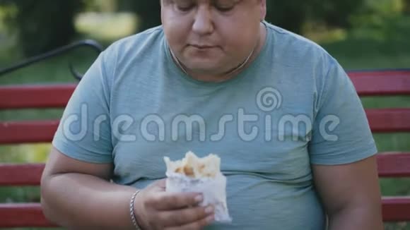 一个胖子在公园的长凳上吃有害的快餐喝咖啡视频的预览图