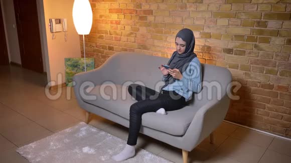 在舒适的家中年轻迷人的穆斯林女性戴着头巾在沙发上放松手机的特写镜头视频的预览图