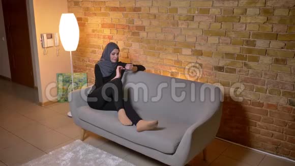 睡在沙发上悠闲地坐着的穆斯林成年女性戴着头巾上网的特写照片视频的预览图