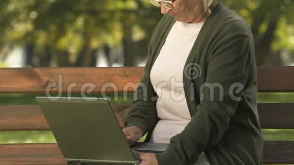 老年女性输入笔记本电脑键盘坐在长凳上上网搜索应用程序视频的预览图