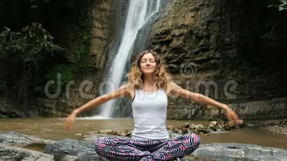 女人坐在莲花的位置双手折叠在一个巨大的瀑布上的名字姿势视频的预览图