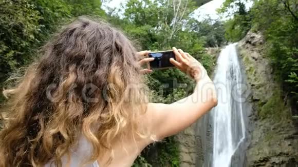 女人把注意力放在智能手机屏幕上拍一张公园瀑布的照片视频的预览图