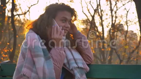 在秋日公园白种人卷发女人坐在长凳上愉快地用手机聊天的肖像视频的预览图