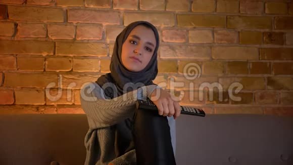 年轻迷人的穆斯林女性戴着头巾坐在电视上一边看电视一边拿着遥控器的特写镜头视频的预览图