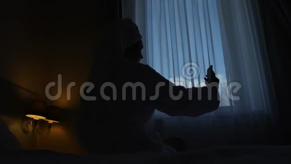 穿浴袍的女孩坐在床上的酒店窗户前视频的预览图