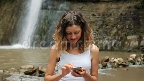 女人在大自然中使用智能手机背景是一个大瀑布依赖互联网视频的预览图