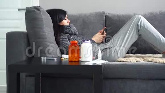 生病的女人在家里躺在沙发上用智能手机视频的预览图