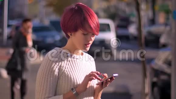 漂亮的粉红色头发女孩的肖像带着智能手机在模糊的街道背景下转向相机视频的预览图