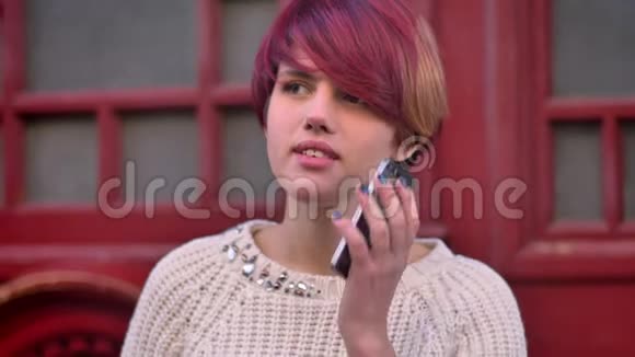 年轻的白种人粉红色头发的女孩在红色的门背景上愉快地在智能手机上交谈的肖像视频的预览图