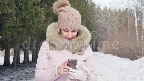一个快乐的女人在雪地冬天的森林里散步时在她的智能手机上写着一条信息的肖像慢动作视频的预览图
