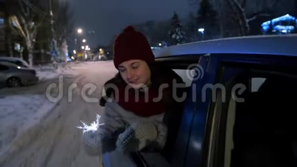 年轻快乐的女人站在车窗外在下雪的冬夜玩着火花漂亮女孩开车很有趣视频的预览图