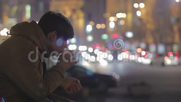 年轻孤独的人在街上喝着热茶看着夜城悲伤视频的预览图