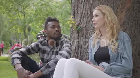 公园里坐在一棵老树下谈天说地的金发美女和美籍黑人帅哥视频的预览图