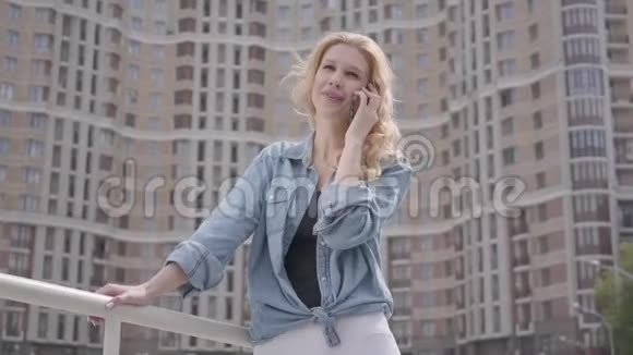 自信微笑的金发女人穿着牛仔裤夹克在摩天大楼前用手机说话城市生活方式女性城市视频的预览图