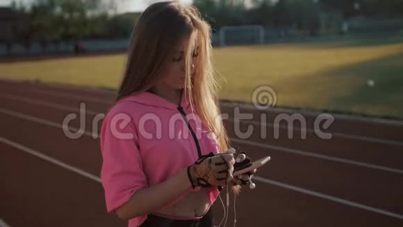 跑步者在红色跑步轨道上听智能手机音乐应用程序带着耳机和智能手机的女运动员视频的预览图