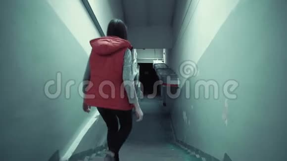 年轻的黑发女人穿着红色无袖夹克在黑暗中走着长长的楼梯视频的预览图