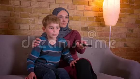 小男孩和他的穆斯林母亲戴着头巾坐在沙发上看电视的肖像视频的预览图