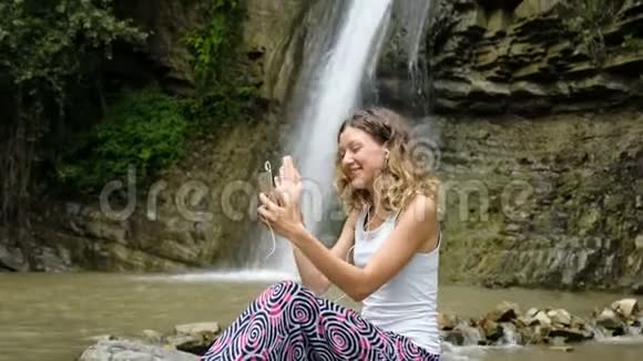 在大瀑布的背景下女人通过手机上的视频通话在耳机中进行交流视频的预览图