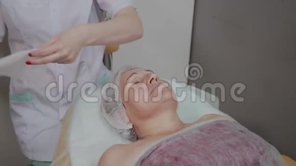 专业美容师在美容办公室对一位老年妇女的脸上施加擦拭视频的预览图