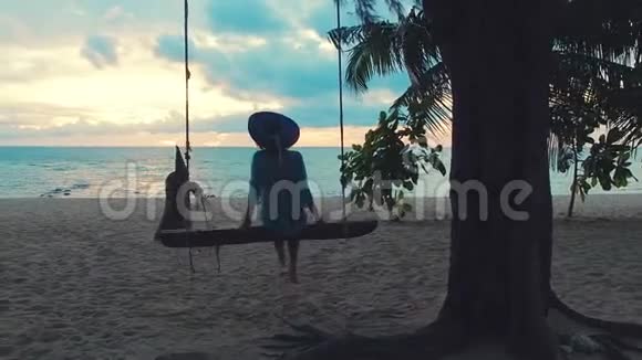日落时戴着帽子的漂亮女孩在海边荡秋千坐在绞刑架上的漂亮年轻女人视频的预览图