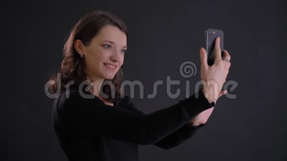 年轻迷人的白种人长发女孩微笑着用智能手机在黑色上制作自拍照片的肖像视频的预览图