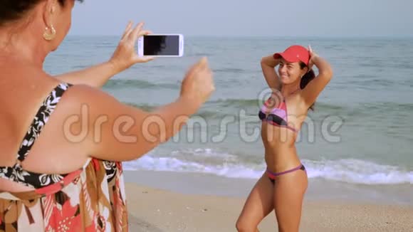 成熟的女人为她晒黑的女儿在海边的泳装上智能手机拍照视频的预览图