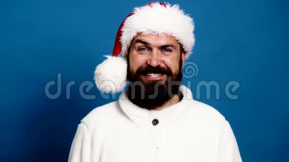 戴着一顶红色新年帽子的留胡子的人眉毛一动脸上带着蓝色的背景微笑有趣的圣诞老人概念视频的预览图