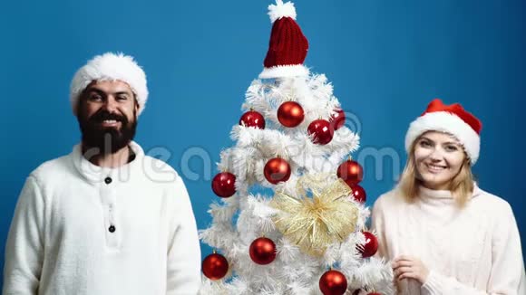 戴着新年帽子的大胡子男人和女孩在蓝色背景下的圣诞树附近表现出不同的情绪爱视频的预览图