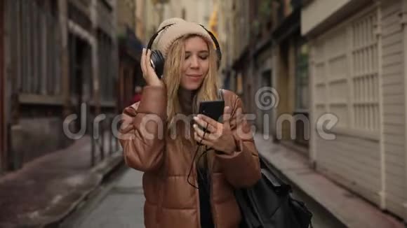 笑着笑着英俊的年轻女子戴着耳机听着智能手机里的音乐在街上跳舞视频的预览图