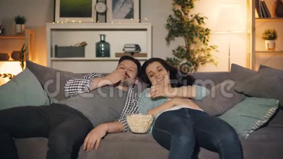 快乐的年轻夫妇在电视上看滑稽节目笑着在家吃爆米花视频的预览图