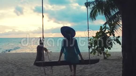 日落时戴着帽子的漂亮女孩在海边荡秋千坐在绞刑架上的漂亮年轻女人视频的预览图