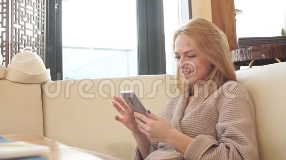 美丽的金发女人在浴室的休息区休息看着智能手机上的照片视频的预览图
