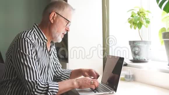 有胡子的成熟男人在家拿着笔记本电脑工作坐在靠近窗户的书桌上视频的预览图