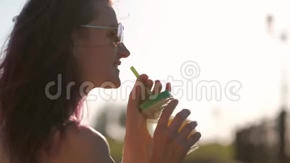 戴着太阳镜的年轻美女正在城市街道上用吸管从玻璃罐里喝鸡尾酒视频的预览图
