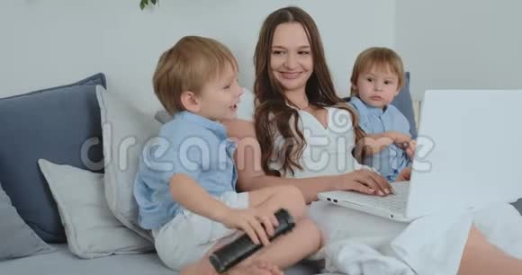 妈妈坐在沙发上拿着笔记本电脑两个男孩的孩子唱歌亲吻他们的妈妈视频的预览图