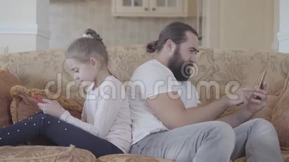 可爱的女儿和她的父亲坐在客厅的沙发上带着智能手机在舒适的日子里不寒而栗父亲和视频的预览图
