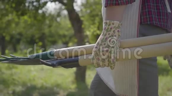 戴着花园手套的农夫手里拿着一把铲子和一把干草叉在花园里走来走去农村概念视频的预览图