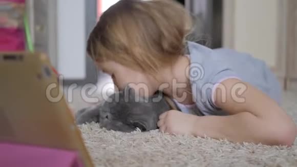 可爱的小女孩拿着数字平板电脑抱着你最喜欢的猫躺在地板上美丽的女孩在玩视频的预览图