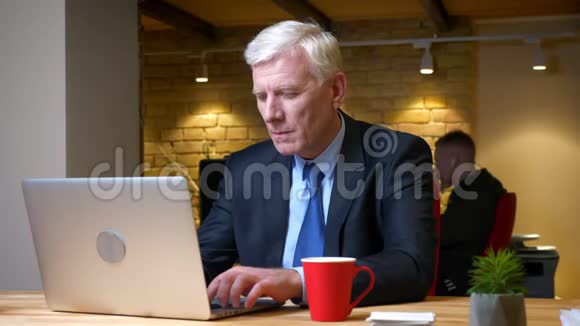 一个老白种人商人在办公室里用手提电脑在办公室里喝咖啡的特写镜头视频的预览图