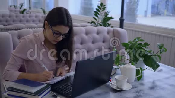 远程在线学习戴眼镜的快乐年轻女子的画像在坐在桌旁的笔记本上写着笔记视频的预览图