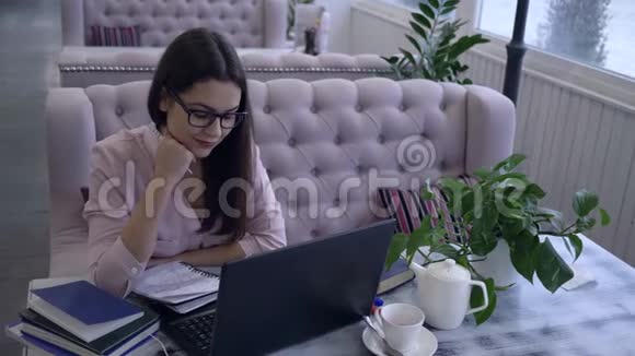 现代教育技术戴眼镜的漂亮女孩用笔记本电脑用笔写笔记视频的预览图
