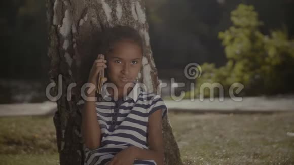 快乐的小女孩在树下用手机说话视频的预览图