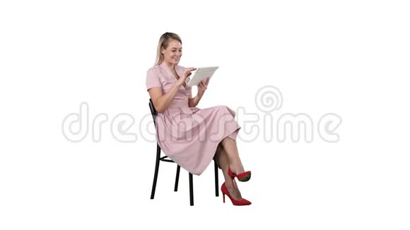 有趣的漂亮年轻女人穿着粉红色的连衣裙坐在椅子上在白色背景上使用平板电脑视频的预览图